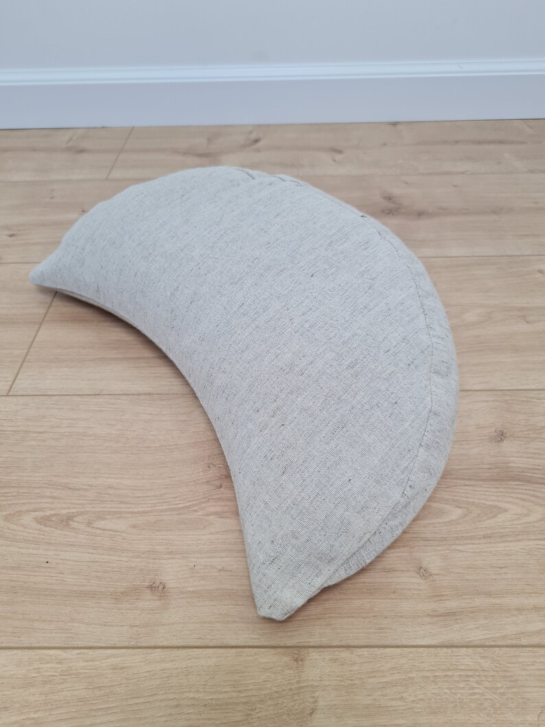 May include: A crescent-shaped pillow made of a light gray linen fabric. The pillow is lying on a light brown wooden floor.