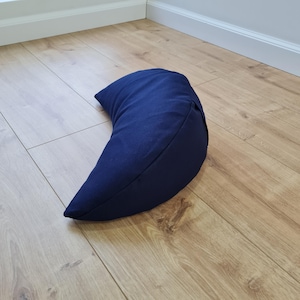 May include: A crescent-shaped meditation cushion in a solid dark navy blue color. The pillow features a smooth, fabric-covered exterior with a curved design that tapers to pointed ends, providing a firm and structured shape for support.