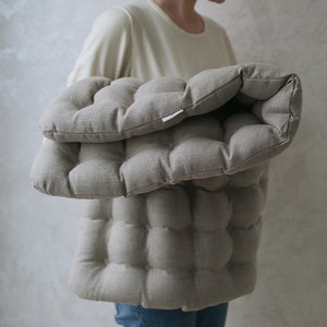 May include: A beige quilted cushion with a textured fabric. The cushion is folded in half and held by a person.