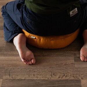 May include: A person sits cross-legged on a yellow meditation cushion with a zipper detail. The cushion is on a wood floor.