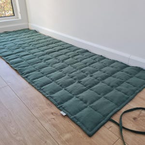 Puede incluir: Una alfombrilla rectangular acolchada de color verde bosque con un patrón de costuras en forma de cuadrícula en toda su superficie. La alfombrilla incluye largas cintas de tela a juego en un extremo para enrollarla y asegurarla. Una pequeña etiqueta blanca es visible en el borde.