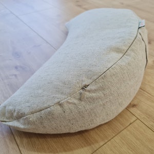 May include: A crescent-shaped meditation cushion with a zipper closure. The cushion is made of a light beige fabric with a subtle texture.