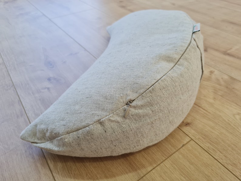 May include: A crescent-shaped meditation cushion with a zipper closure. The cushion is made of a light beige fabric with a subtle texture.