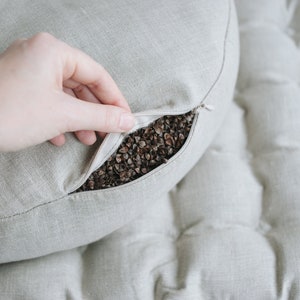 May include: A light gray linen cushion with a zipper opening, revealing a filling of buckwheat hulls.