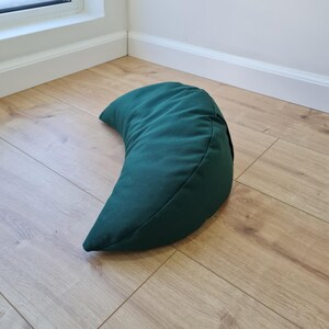 May include: A crescent moon-shaped meditation cushion in a solid dark forest green color. The pillow features a smooth, matte fabric exterior with visible stitched seams along the curved edges, creating a firm and structured half-moon silhouette.