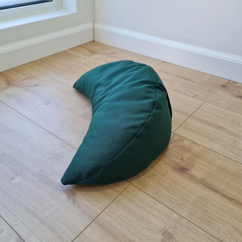 May include: A crescent moon-shaped meditation cushion in a solid dark forest green color. The pillow features a smooth, matte fabric exterior with visible stitched seams along the curved edges, creating a firm and structured half-moon silhouette.