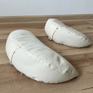 May include: Two off-white crescent-shaped meditation cushions on a light brown wooden floor.