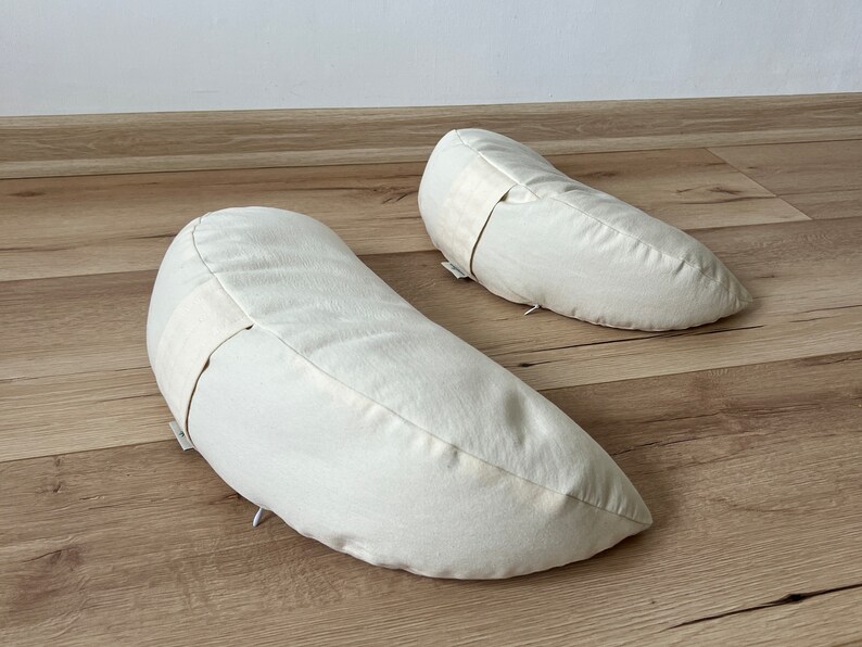 May include: Two off-white crescent-shaped meditation cushions on a light brown wooden floor.