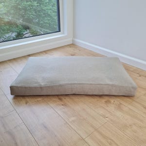 Puede incluir: Un cojín rectangular para mascotas con una funda de tela de color beige claro texturizado. El cojín tiene un diseño plano y de perfil bajo con bordes cosidos limpios, proporcionando una superficie de descanso sencilla y funcional.