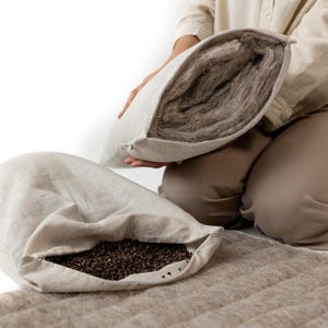 May include: A person kneels, holding a light-colored pillow open to reveal the filling. Another pillow and a rectangular mat are visible. The pillow on the left is open, showing dark brown grains. The mat and pillows are a neutral color.