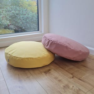 Könnte beinhalten: Zwei runde Bodenkissen, eines in Senfgelb und das andere in Altrosa. Beide Kissen haben eine glatte, matte Stoffoberfläche und ein schlichtes, kreisförmiges Design mit einem kleinen seitlichen Etikett am gelben Kissen.