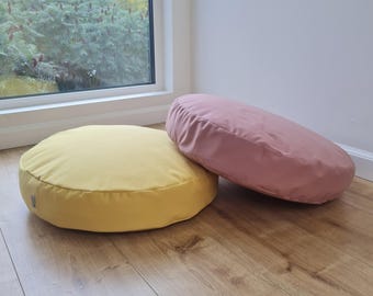 Könnte beinhalten: Zwei runde Bodenkissen, eines in Senfgelb und das andere in Altrosa. Beide Kissen haben eine glatte, matte Stoffoberfläche und ein schlichtes, kreisförmiges Design mit einem kleinen seitlichen Etikett am gelben Kissen.