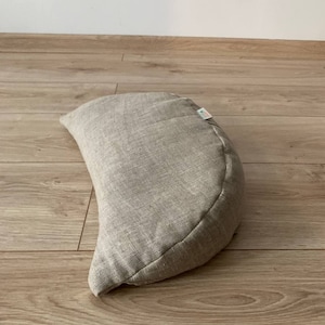 May include: A crescent-shaped meditation cushion made of natural linen fabric. The cushion is light beige in color and has a simple, understated design.