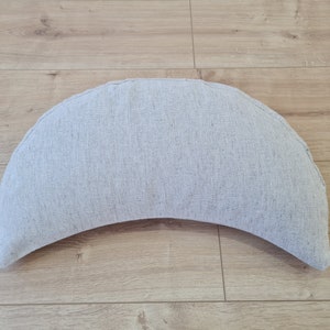May include: A crescent-shaped cushion made of a light gray fabric. The cushion is sitting on a wooden floor.