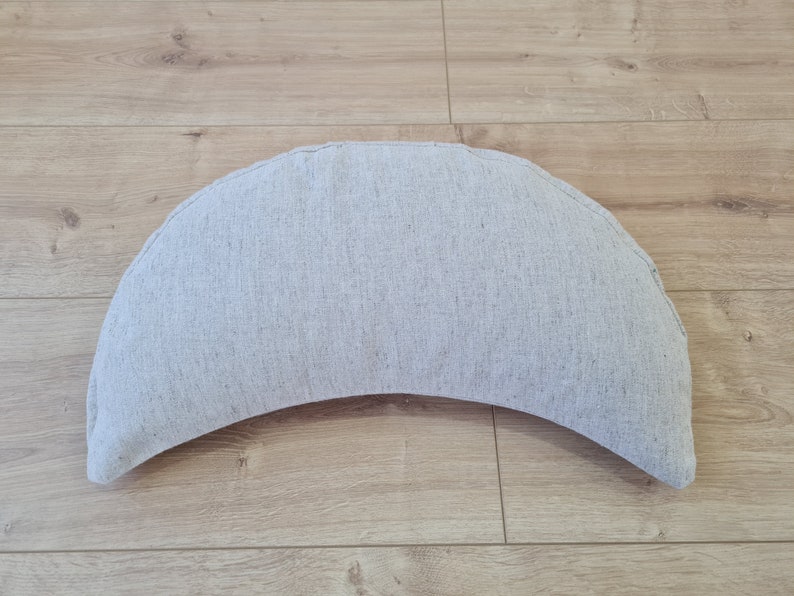 May include: A crescent-shaped cushion made of a light gray fabric. The cushion is sitting on a wooden floor.