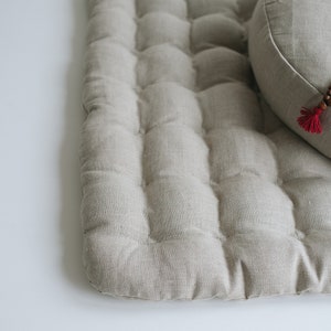 May include: A light gray meditation cushion with a tufted surface and a round cushion with a beaded necklace on top.