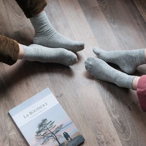 May include: Two people wearing gray socks sit on a wood floor with a magazine titled "LA BOUSSOLE" in front of them.