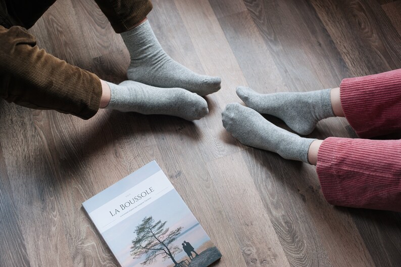 May include: Two people wearing gray socks sit on a wood floor with a magazine titled "LA BOUSSOLE" in front of them.