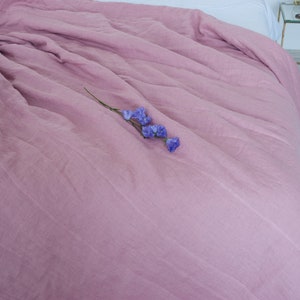 May include: A dusty rose-colored linen duvet cover draped over a bed. The bedding has a soft, textured appearance. A sprig of purple flowers rests on the duvet. The bed is next to a small glass and gold side table.