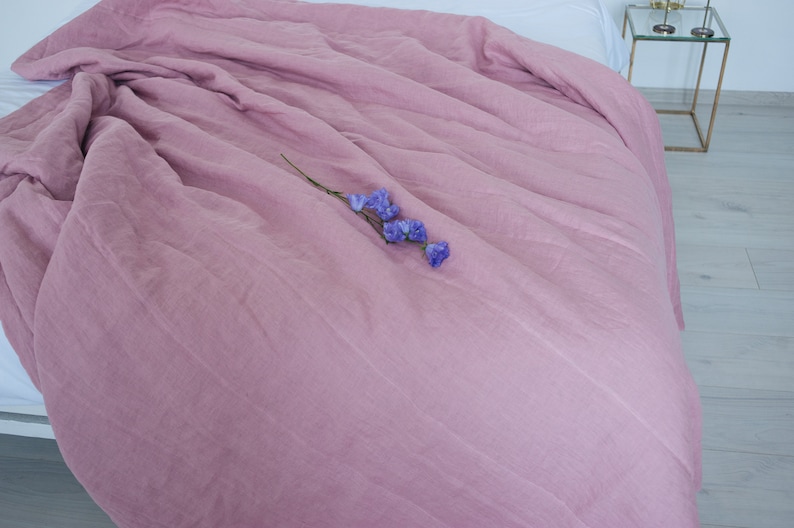 May include: A dusty rose-colored linen duvet cover draped over a bed. The bedding has a soft, textured appearance. A sprig of purple flowers rests on the duvet. The bed is next to a small glass and gold side table.