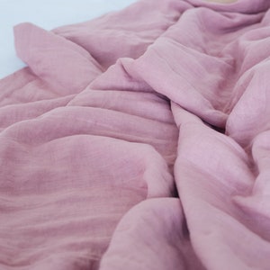 May include: A close-up shot of a soft, dusty rose-colored blanket. The fabric is wrinkled and draped, creating shadows and texture. The blanket appears to be made of a lightweight material, possibly linen or cotton.
