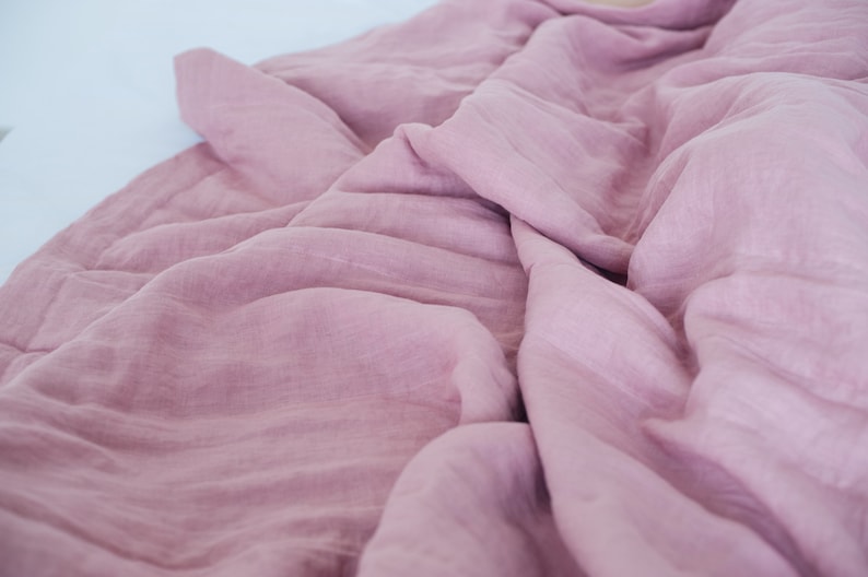 May include: A close-up shot of a soft, dusty rose-colored blanket. The fabric is wrinkled and draped, creating shadows and texture. The blanket appears to be made of a lightweight material, possibly linen or cotton.