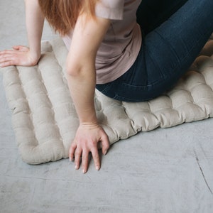 May include: A beige linen cushion with a quilted design. The cushion is on a light gray floor.