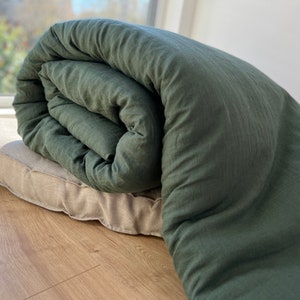 Könnte beinhalten: Eine zusammengerollte, dunkelgrüne Bettdecke liegt auf einem beigen rechteckigen Kissen. Die Bettdecke besteht aus einem strukturierten Stoff, und das Kissen hat ein schlichtes, neutrales Design. Die Artikel befinden sich auf einem Holzboden in der Nähe eines Fensters.