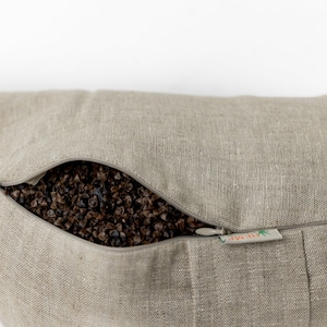 May include: A beige linen pillow with a zipper opening revealing a buckwheat hull filling. The pillow has a small label that reads "All-Natural".