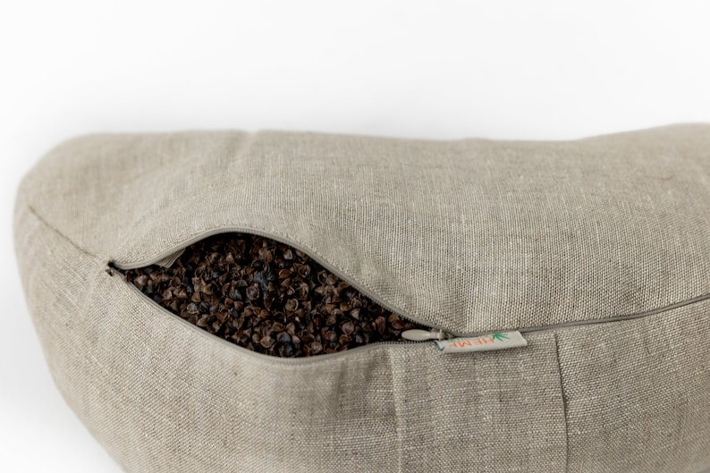 May include: A beige linen pillow with a zipper opening revealing a buckwheat hull filling. The pillow has a small label that reads "All-Natural".