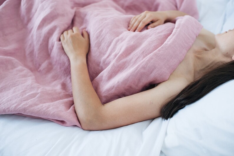 May include: A person is lying in bed, covered by a soft, dusty rose-colored linen blanket. The bedsheets are white, creating a contrast with the blanket. The scene evokes a sense of comfort and relaxation.