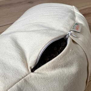 May include: An off-white, cylindrical bolster pillow made of textured fabric. A white zipper is partially open, revealing dark brown buckwheat hulls inside. A small fabric tag attached to the zipper reads HEMP and includes a green leaf icon.