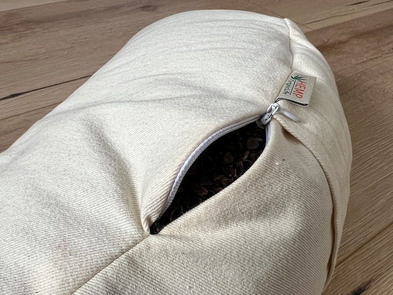 May include: An off-white, cylindrical bolster pillow made of textured fabric. A white zipper is partially open, revealing dark brown buckwheat hulls inside. A small fabric tag attached to the zipper reads HEMP and includes a green leaf icon.