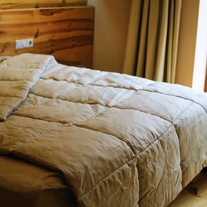 May include: A close-up of a beige duvet with a quilted pattern, lying on a bed with a wooden headboard. The duvet is folded back to reveal the white sheets underneath.