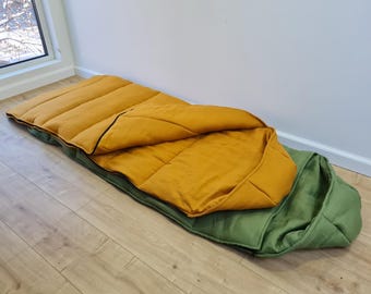 Può includere: Un sacco a pelo aperto, giallo senape e verde oliva. L'interno è trapuntato in giallo senape, mentre l'esterno è verde oliva. Il sacco a pelo è appoggiato su un pavimento in legno. Ideale per il campeggio.
