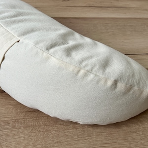 May include: A white crescent-shaped meditation cushion with a white fabric strap. The cushion is made of natural materials and is designed for comfort and support during meditation.