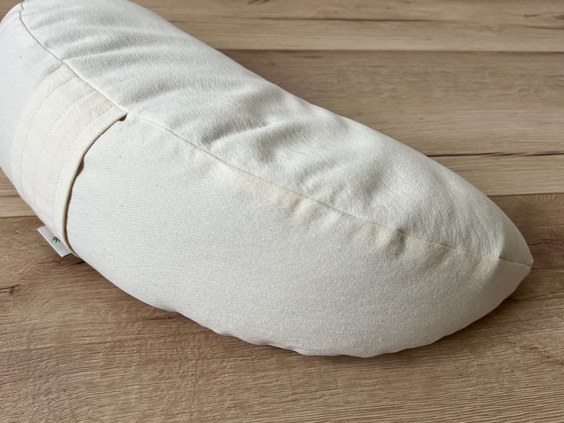 May include: A white crescent-shaped meditation cushion with a white fabric strap. The cushion is made of natural materials and is designed for comfort and support during meditation.
