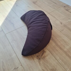 May include: A crescent-shaped pillow in a solid dark brown color. The pillow features a smooth fabric exterior with a visible seam running along the curved edge, creating a distinct half-moon silhouette that rests on a flat surface.