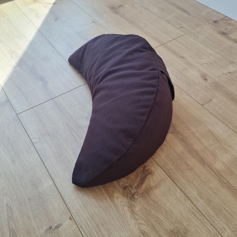 May include: A crescent-shaped pillow in a solid dark brown color. The pillow features a smooth fabric exterior with a visible seam running along the curved edge, creating a distinct half-moon silhouette that rests on a flat surface.