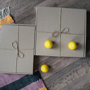 May include: Two brown cardboard boxes tied with twine, one on top of the other, with three yellow candles on the top box. The boxes are on a wooden surface with a striped rug.