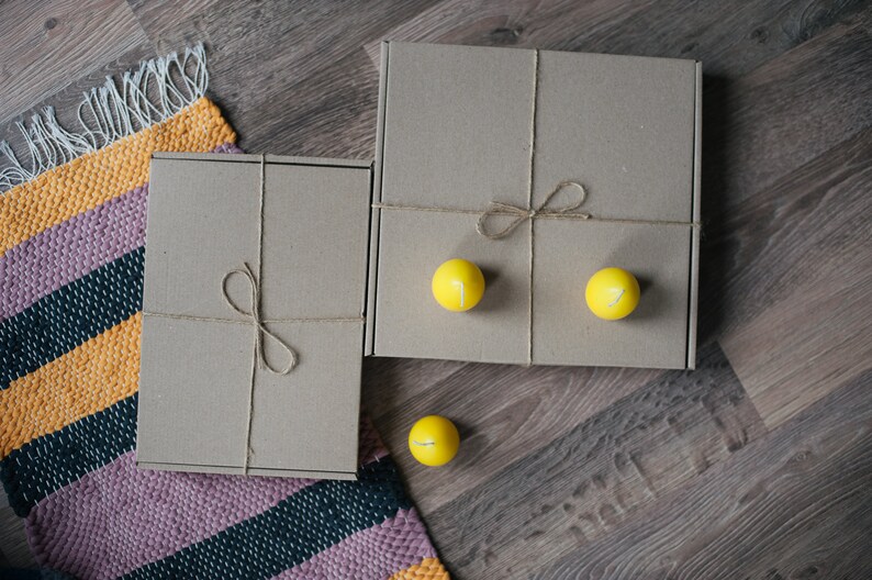 May include: Two brown cardboard boxes tied with twine, one on top of the other, with three yellow candles on the top box. The boxes are on a wooden surface with a striped rug.