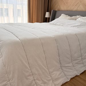 Könnte beinhalten: Eine weiße, gesteppte Bettdecke bedeckt ein Bett mit passendem Kissen und einem grauen Kopfteil. Die Bettdecke hat vertikale Steppnähte und ein weiches, einladendes Aussehen. Der Raum hat eine neutrale Farbpalette.