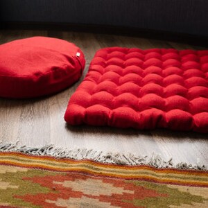 May include: A red round meditation cushion and a red rectangular meditation cushion with a quilted surface. Both cushions are on a wooden floor with a patterned rug in the foreground.