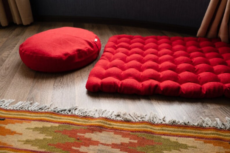 May include: A red round meditation cushion and a red rectangular meditation cushion with a quilted surface. Both cushions are on a wooden floor with a patterned rug in the foreground.