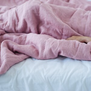 May include: A bed covered with a soft, dusty rose-colored linen duvet. The duvet is draped over a white bed sheet. A person's arm is visible, resting under the duvet. The bedding creates a cozy and inviting atmosphere.