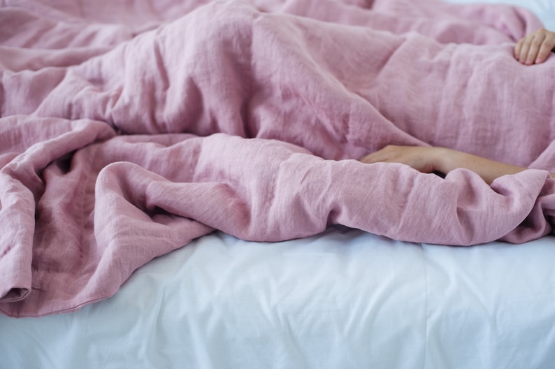 May include: A bed covered with a soft, dusty rose-colored linen duvet. The duvet is draped over a white bed sheet. A person's arm is visible, resting under the duvet. The bedding creates a cozy and inviting atmosphere.