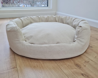 Puede incluir: Una cama redonda para mascotas de color beige, con un borde acolchado elevado. La cama está hecha de una tela texturizada y se encuentra sobre un suelo de madera. El cojín central es de un beige más claro.