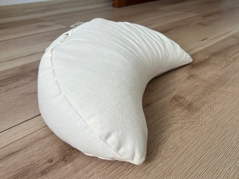 May include: A crescent-shaped meditation cushion in an off-white, natural canvas fabric. The pillow features a curved design with visible stitched seams along the edges and a small fabric tag attached to the top side of the cushion.