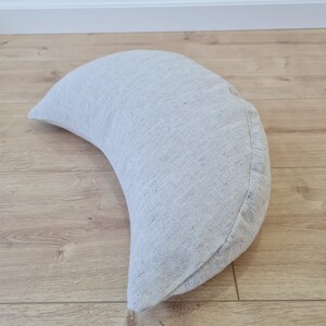 May include: A crescent-shaped meditation cushion made of a light gray fabric with a subtle texture.