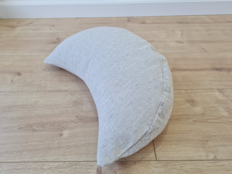 May include: A crescent-shaped meditation cushion made of a light gray fabric with a subtle texture.